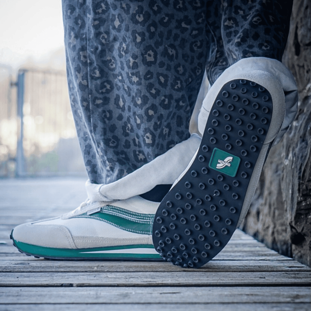 Close-up of a person wearing white sneakers with green accents on a wooden floor.