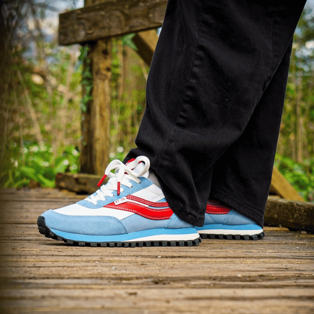 Blue, white, and red trainers worn with black pants on a wooden bridge.