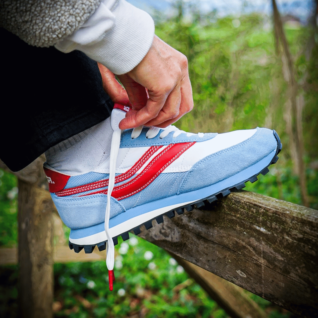 Person tying shoe laces with red metal anklets on a blue, white, and red sneaker outdoors.
